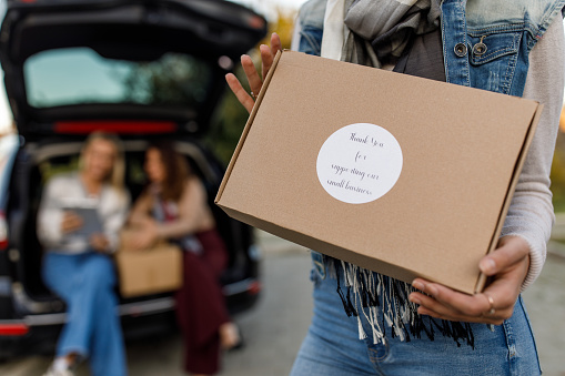 Midsection of unrecognizable young woman standing in front of an open car trunk with two of her colleagues, holding a package  labeled ''Thank you for supporting our small business''.