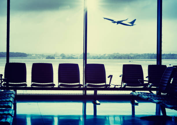 Side view of a commercial airplane with a clear reflection on its fuselage. Passengers are boarding the aircraft via external stairs. The airport terminal is visible in the background under a dramatic evening sky, capturing a dynamic moment of air travel.