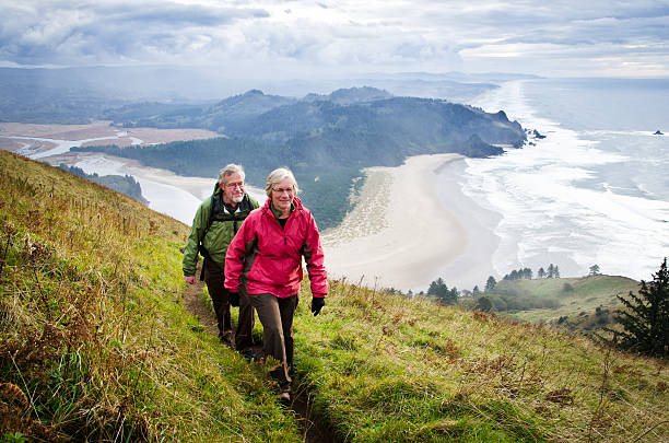 A senior couple hikes on a trail far above a rugged coastline on a stormy day. Cascade Head, Oregon. The Salmon River estuary is seen at left.
