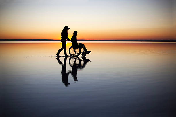 dependence helpful woman takes care of the disabled man in front of the beautiful lake sunset. mid distance stock pictures, royalty-free photos & images