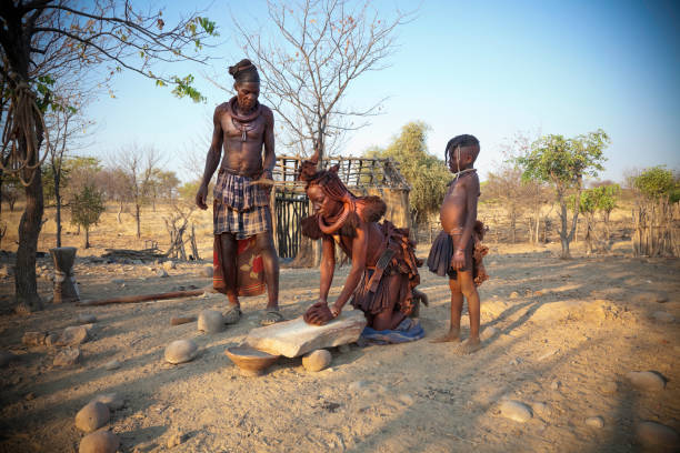 Himba family The mother is grinding maize with a stone to make flour. There is some motion blur of the woman's head and hands as she works. kaokoveld stock pictures, royalty-free photos & images