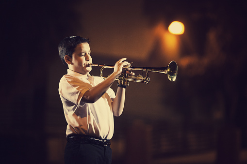 eleven years old kid playing the trumpet outdoors with a spotlight from a street lamp in the back