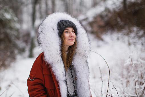 Redhead woman in red jacket with fur and hat in winter forest. Pretty woman enjoying vacation. Girl in hood in nature. Feel happiness. Hoarfrost and snow on trees. Holidays in mountains