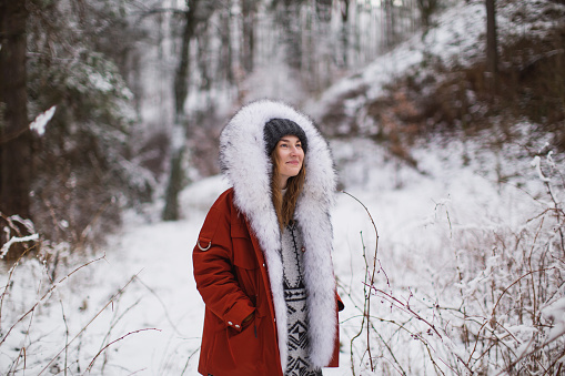 Redhead woman in red jacket with fur and hat in winter forest. Pretty woman enjoying vacation. Girl in hood in nature. Feel happiness. Hoarfrost and snow on trees. Holidays in mountains