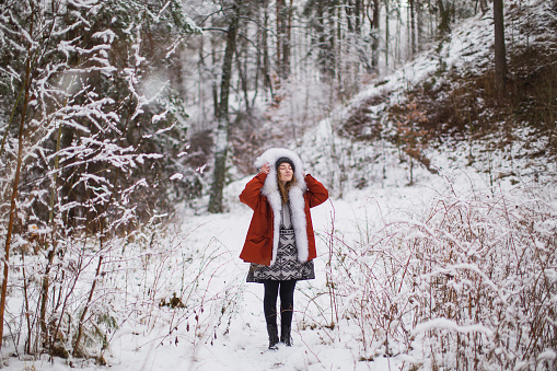 Redhead woman in red jacket with fur and hat in winter forest. Pretty woman enjoying vacation. Girl in hood in nature. Feel happiness. Hoarfrost and snow on trees. Holidays in mountains