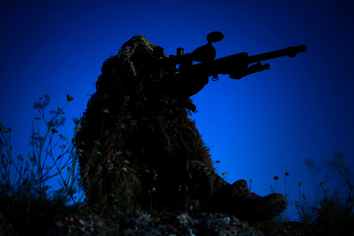 special operation forces sniper in ghillie (camouflage) suit with a high-precision rifle with optic scope in a combat position in the mountains at night