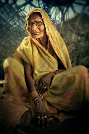 Bespectacled, senior Indian woman sitting on ground, she is wearing traditional Indian dress and golden jewelry, photo shot in her village.