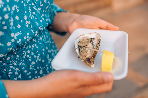 Close up female hand holding take away food tray with fresh opened oyster and lemon slices at street food market, festival, event. Selective focus. Valencia, Spain, Europe.