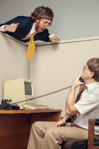 A manager in full suit leans over an office cubicle and yells at his co-worker, looking very angry.  The other business man is on a phone call and stares at the yelling man.  1980's styling, complete with vintage computer.  Vertical.