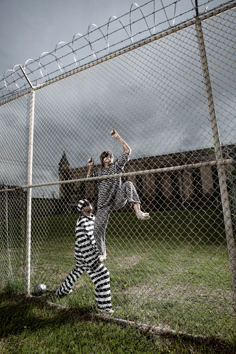 A male inmate attached to a ball and chain assists a female prisoner in a daring escape attempt, climbing over a tall prison fence.