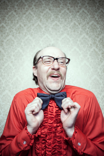 A balding man in a retro frilly red tuxedo shirt, mustache, and glasses, makes a goofy face while straightening his bow tie.  He looks very happy and proud of himself.  Vintage damask pattern wallpaper on wall in the background.  Vertical with copy space.
