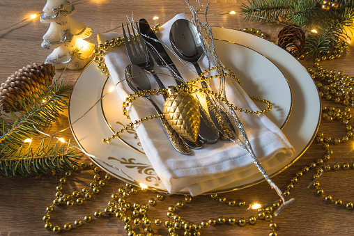 Fork, knife, spoons on white linen napkin and white plates with golden decoration and christmas lights