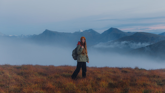 Beautiful female with a backpack in yellow hat admiring the autumn in the mountains in walking in the clouds during twilight in More og Romsdal county
