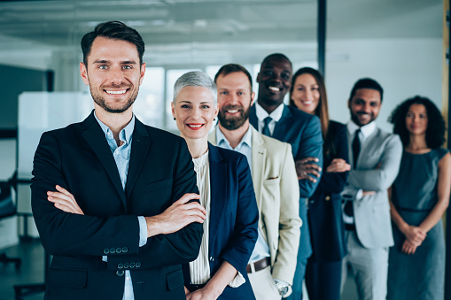 Portrait of handsome smiling businessman with his colleagues. Multi-ethnic group of business persons standing in a row in modern office. Successful team leader and his team in background.