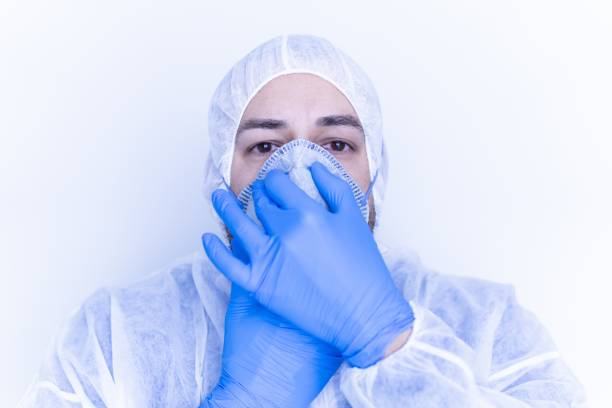 istock Medical worker in a plastic suit putting on a face mask with blue plastic gloves 1439239608