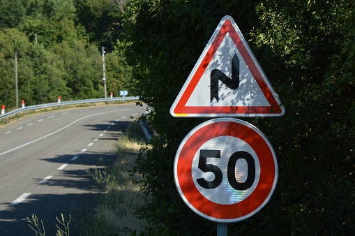 Road traffic hazard warning of double bend and a speed limit 50 sign on the side of a road with trees