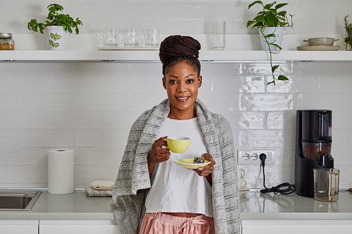 Portrait of a beautiful young adult black woman at the kitchen counter, on a cold winter morning, drinking hot coffee wit a vegan sweet cookie