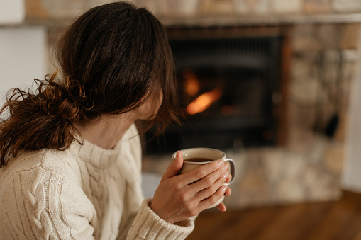 Young woman sitting at home by the fireplace with a hot tea or coffee mug and warming her hands, she is wearing white woollen sweater. Cold houses in Europe concept. Energy crisis.