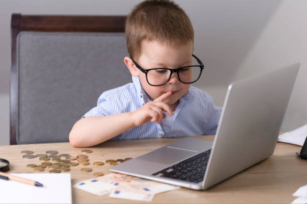 a little boy in glasses sits at a table and looks at a laptop screen. - paying children only retail childhood imagens e fotografias de stock