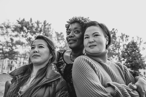 Black-and-white photo of three multiracial friends embracing at the city park looking up.