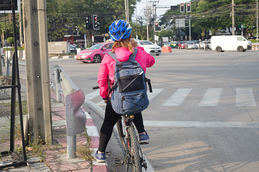 Rear view of cycling adult thai women waiting at junction. Woman is wearing cycling helmet and a backpack and a blue helmet. She has a pink colored jacket. Scene is in Bangkok Nawamin close to Nawamin Road