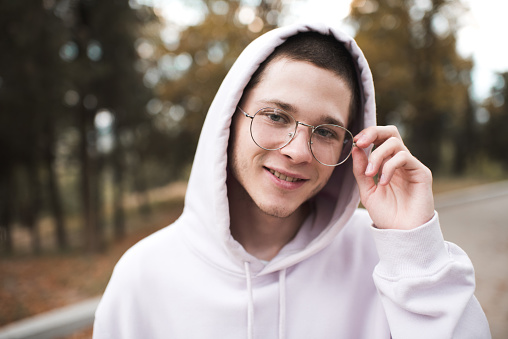 Smiling cute teenager boy 19-20 year old wear glasses and hood over nature background outdoor close up. Happy male kid student.