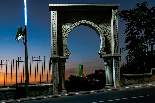 Seen through à sculpted door with Ottoman style arch. Flag pole with Algerian flags and a blue sky with golden hour sunlights.