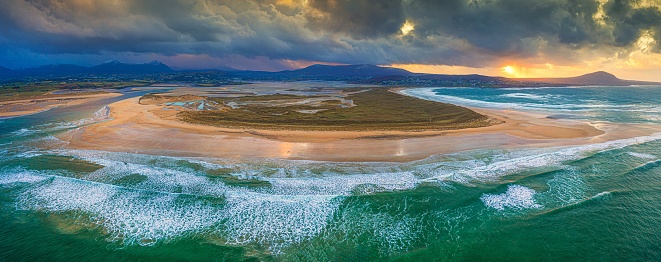 A panoramic aerial shot of land surrounded by the sea under an orange sky at sunset