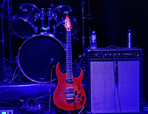 An electric guitar with drums and a big amp in the background under purple lighting conditions