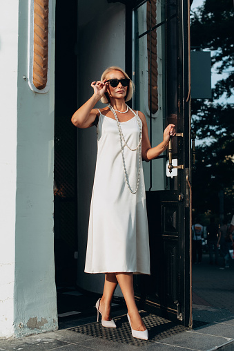full length portrait of a fashionable old woman in white dress and sunglasses exits the door of the building