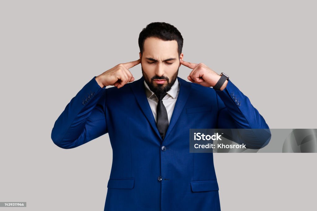 Bearded man covering ears annoyed by unpleasant noise, loud voices, difficult to listen high sound. Portrait of bearded man covering ears annoyed by unpleasant noise, loud voices, difficult to listen high sound, wearing official style suit. Indoor studio shot isolated on gray background. Avoidance Stock Photo