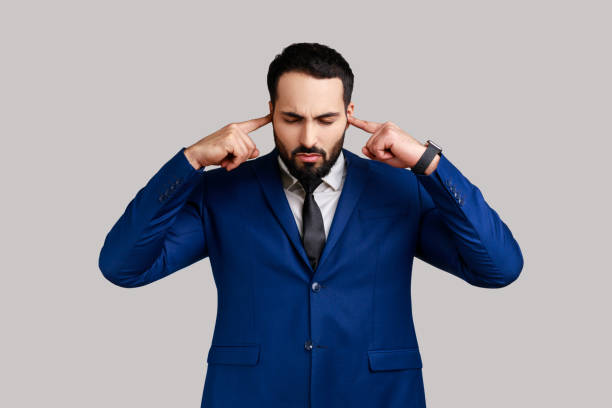 Bearded man covering ears annoyed by unpleasant noise, loud voices, difficult to listen high sound. Portrait of bearded man covering ears annoyed by unpleasant noise, loud voices, difficult to listen high sound, wearing official style suit. Indoor studio shot isolated on gray background. man closing ears photos stock pictures, royalty-free photos & images