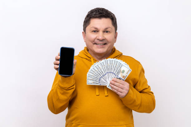 istock Man holding empty screen smartphone ad fan of hundred dollars, looking at camera with smile. 1439312400