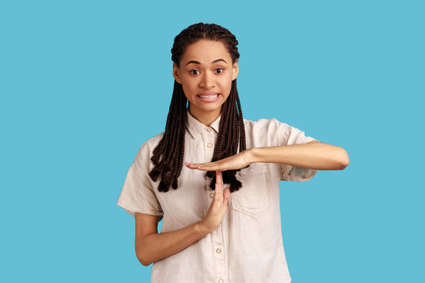 Frustrated woman showing time out gesture, looking with imploring eyes, hurry to meet deadline. I need more time. Portrait of frustrated woman with black dreadlocks showing time out gesture, looking with imploring eyes, hurry to meet deadline. Indoor studio shot isolated on blue background. time out signal stock pictures, royalty-free photos & images