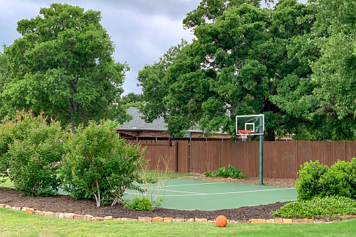 Outdoor shot of backyard of country house in the suburbs with trees, bushes, flowerbed and basketball court, zone for activity at home.