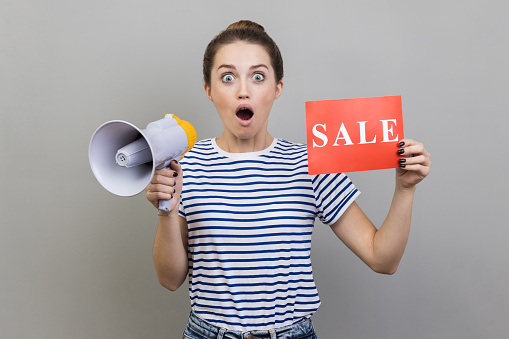 Portrait of shocked astonished amazed woman wearing striped T-shirt holding card with sale inscription and megaphone, making announcement. Indoor studio shot isolated on gray background.