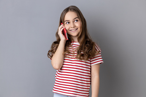 Portrait of satisfied little girl wearing striped T-shirt holding phone near his ear, talking with friends, favorable tariff for calls in roaming. Indoor studio shot isolated on gray background.
