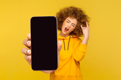 Portrait of crazy positive woman with Afro hairstyle wearing hoodie showing big cell phone display for copy space, showing rock and roll gesture. Indoor studio shot isolated on yellow background.