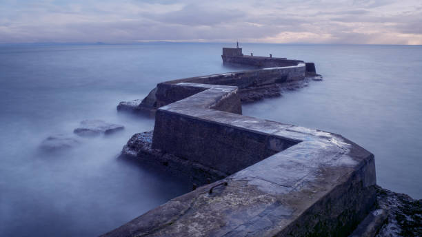 A slightly turbulent sea laps over a concrete wall and steps that lead into the sea, on a sunny day.