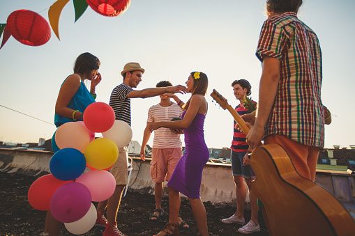 Wide shot of group of friends in colorful outfits hugging each other while bringing balloons and a guitar for a party in a terrace during summer days.