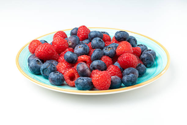 Stock photo showing a close-up, elevated view of a bowlful of cut strawberries, blueberries and bright red, whole raspberries. The strawberry is a popular summer fruit that is considered to be an especially healthy snack, since they are packed with beneficial antioxidants, being a good source of vitamin C. Raspberries contain potassium which assists  heart function, and has been proved to lower blood pressure, whilst omega-3 fatty acids in raspberries prevent stroke and heart disease. They also contain a mineral called manganese, which is necessary for healthy bones and skin and helps regulate blood sugar. Popular with children and adults alike, blueberries have several potential health benefits, including helping to improve memory. Health and wellbeing concept.