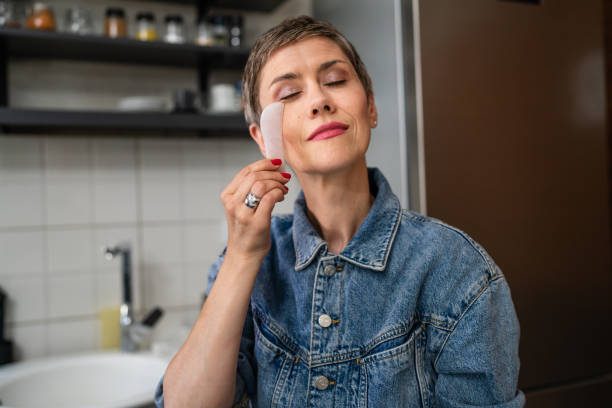 una mujer caucásica madura usando piedra de masaje facial de cuarzo rosa gua sha en la cocina en casa tratamiento de cuidado de la piel relajación y estiramiento facial concepto de rejuvenecimiento de personas reales copian el espacio - quartz caucasian one person energy fotografías e imágenes de stock