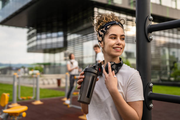 mujer caucásica tomando un freno durante el entrenamiento al aire libre en el gimnasio al aire libre del parque descansando en las barras con agitador de suplementos en la mano agua potable o suplementación sonrisa feliz copia espacio - atleta suplemento fotografías e imágenes de stock