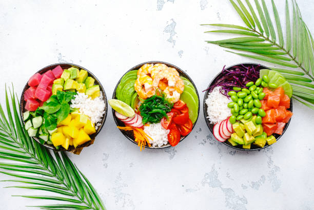 Hawaiian poke bowl set: tuna, salmon, shrimp with avocado, mango, radish, rice and other ingredients. Soy sauce and sesame dressing. White table background, palm leaves, top view stock photo