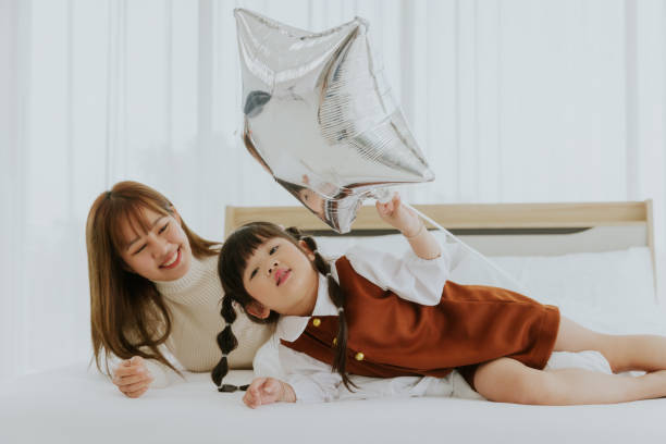 アジアの若い母親と4歳の子供の娘が、白いベッドの上で笑顔で風船を一緒にプレイし、幸せな素敵な家族のコンセプト - 20 25 years adult bed bedroom ストックフォトと画像