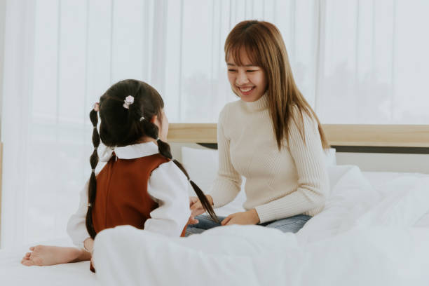 アジアの若い母親と4歳の子供の娘が笑顔で横たわり、白いベッドで一緒にキャンディーを食べて遊ぶ、幸せな素敵な家族のコンセプト - 20 25 years adult bed bedroom ストックフォトと画像