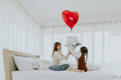Young asian mom smiling holding heart balloon playing with little 4years old kid daughter in bed room, Mother's day concept