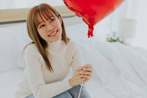 Portrait young pretty asian female smiling holding red heart balloon sitting on bed in bedroom, Love valentine concept