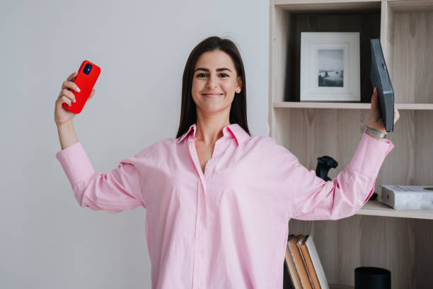 istock I did it! Happy young brunette holding  phone and diary with smile expression dressed in pink shirt against book shelves bending hands showing biceps.Young woman received great news, winner expression 1439377310