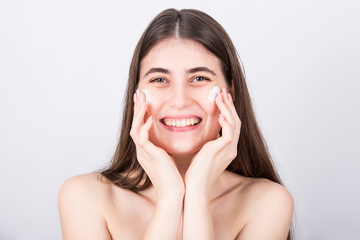 Happy young woman spreads moisturizing face cream on her cheeks. Beautiful girl apply softening moisturizer, isolated on white background. Skincare, cosmetics, beauty spa procedures concept